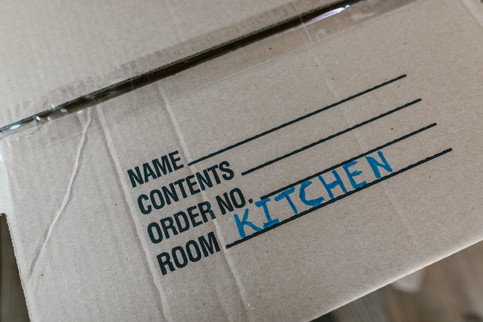 Inside a residential property during a home relocation process, a stack of cardboard moving boxes of various sizes is arranged on a tiled floor near a doorway. Some boxes are sealed with packing tape,